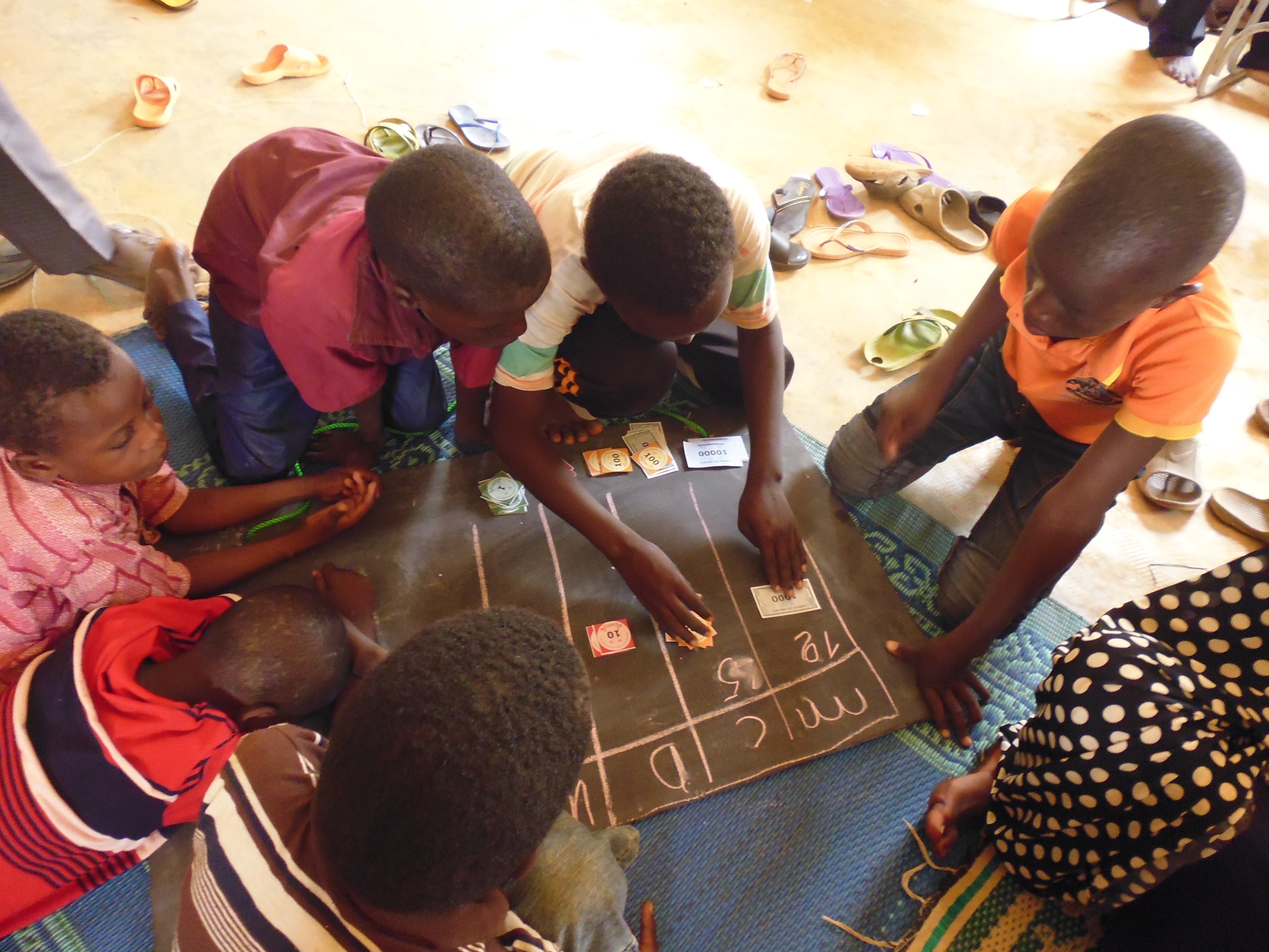 Seven children sit on the floor in a circle, with a chalkboard on the floor in the middle of them. One child places play money notes onto the board while the other children watch during a Teaching at the Right Level-integrated PMAQ programme in a classroom in Niger.